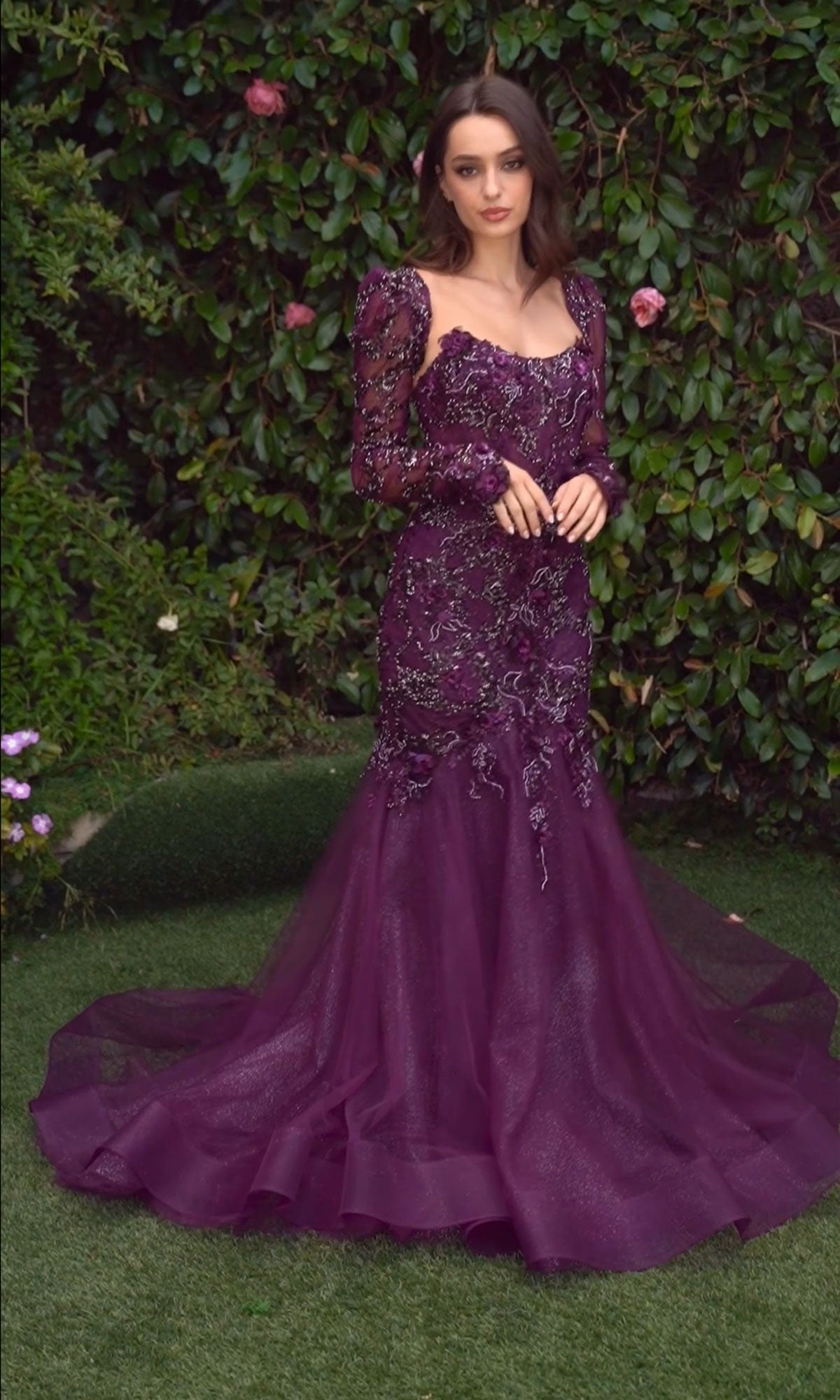 A woman stands outdoors before greenery, wearing the Andrea & Leo MA101 long sleeve formal dress featuring beaded lace, intricate embellishments, and a mermaid skirt flowing elegantly to the ground.